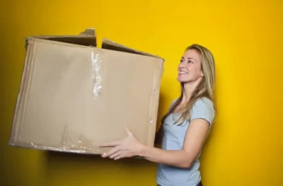 A happy woman carrying a box and smiling in front of a yellow wall